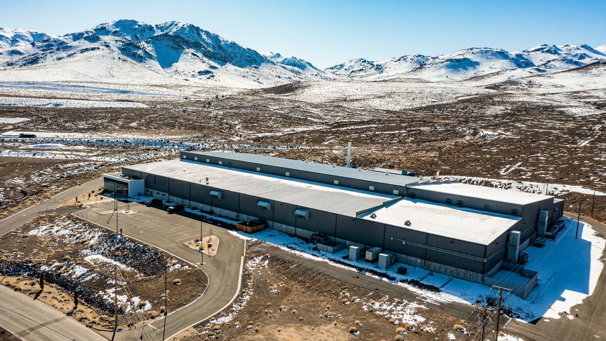 The American Battery Technology Company facility in McCarran, Nevada, with a parking lot at the front, and a road next to the facility under the sun. Snow-covered mountains are in the background.