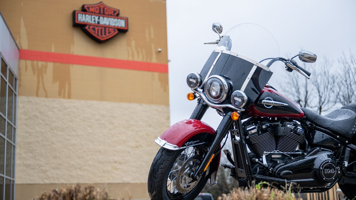 A motorcycle on the bottom right with a tan building with an orange and black Harley-Davidson logo blurred in the background on a cloudy day.