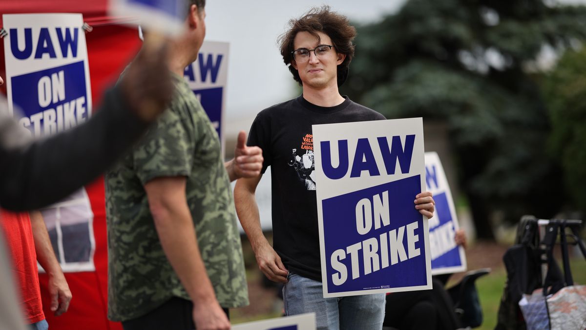 Two autoworkers look at each other with "UAW on Strike" signs in their hands and plastered on makeshift tents.