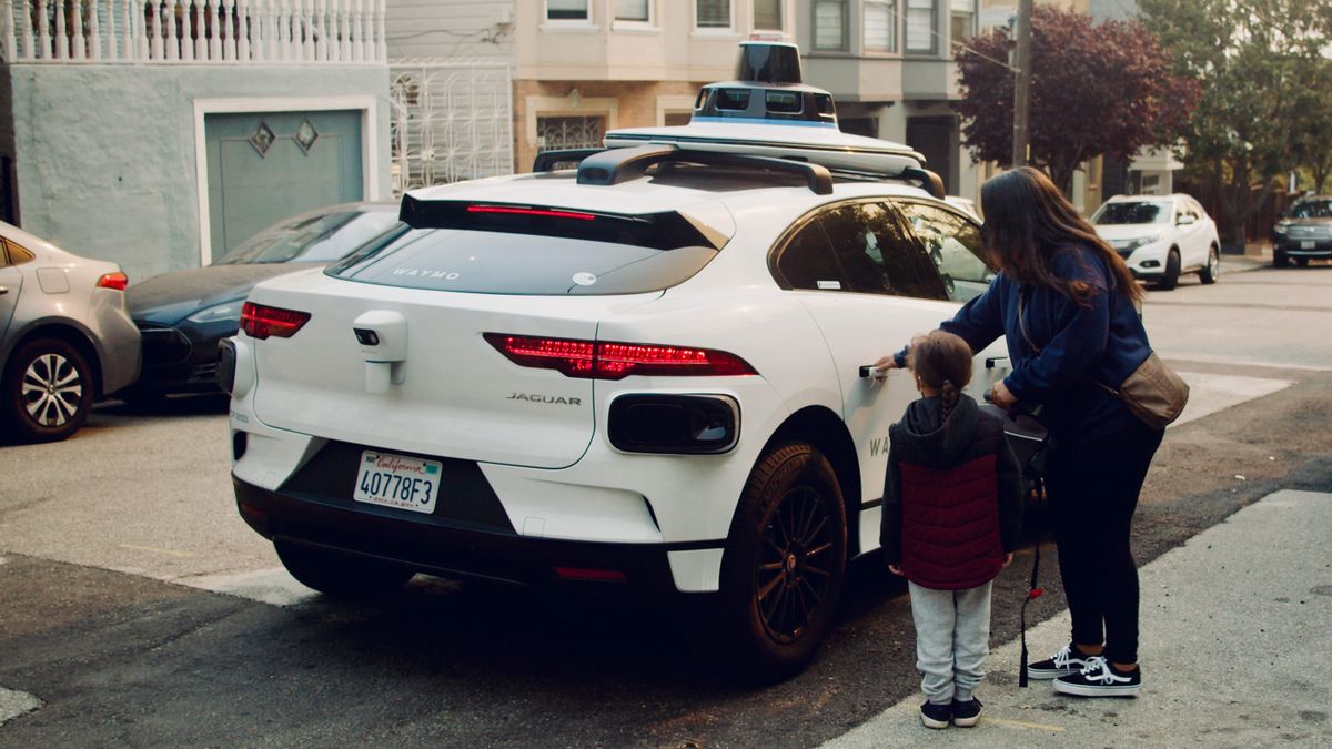 A woman and child are about to enter a white self-driving, ride-hailing vehicle with large sensors on the roof in San Francisco.